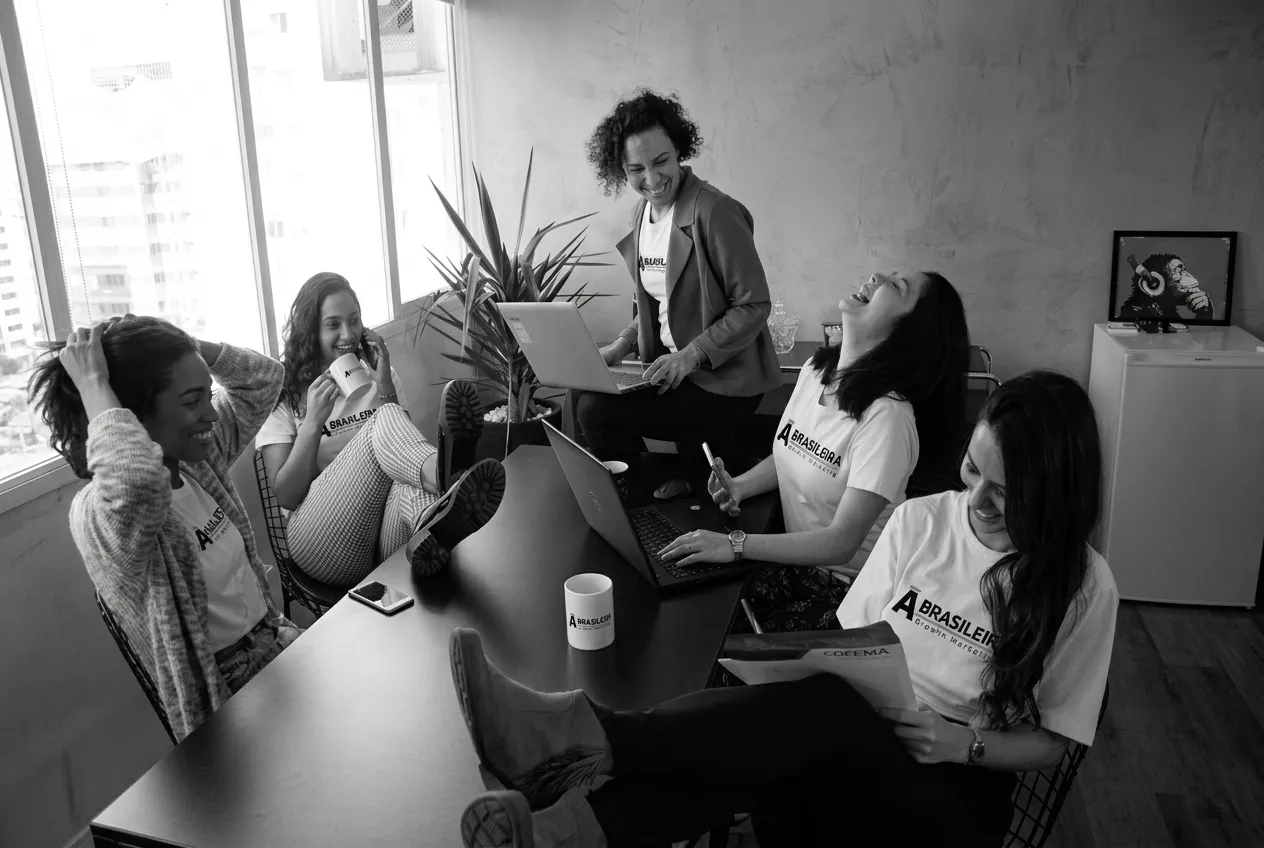 Equipe da À Brasileira reunida em uma mesa de trabalho, conversando entre laptops e canecas da marca